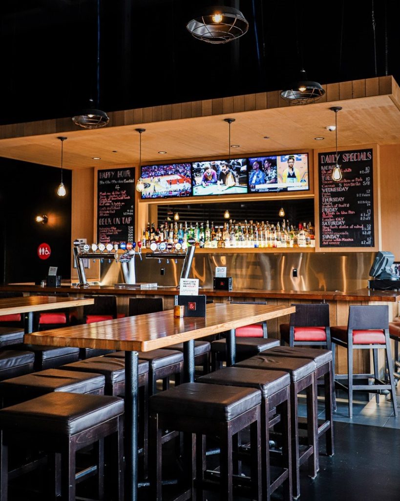 Pub interior with long high tables, leather stools, and flat screens above a stocked bar. above a